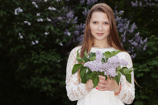 A beautiful girl in a white dress with long hair against the background of a spring lilac bush.