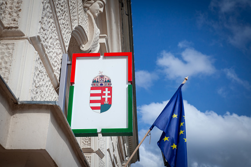 Picture of the coat of arms of hungary, magyarorszag cimere, in front of a flag of the eu, european union, in Pecs, Hungary. The coat of arms of Hungary was adopted on 3 July 1990, after the end of communist rule. The arms have been used before, both with and without the Holy Crown of Hungary, sometimes as part of a larger, more complex coat of arms, and its elements date back to the Middle Ages.