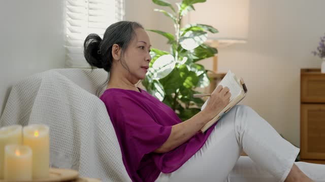 a good looking old female is writing a diary about her beautiful memory happily on a couch in the living room of the house, elderly women is journaling with smiling on her face.
