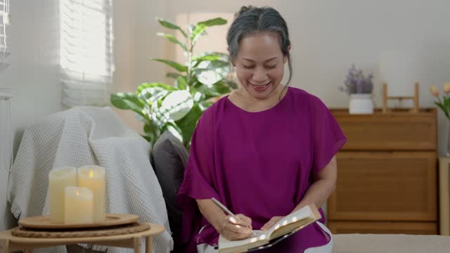 a good looking old female is writing a diary about her beautiful memory happily on a couch in the living room of the house, elderly women is journaling with smiling on her face.