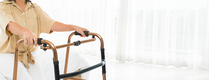 A woman in a tan shirt is using a walker. The walker is made of metal and has a black handle, panorama concept. The woman is sitting on a bed and she is in a relaxed mood.