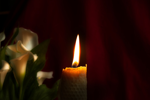 Wax candle with a yellow flame, centrally positioned, burning in a dark environment against a red curtain backdrop that provides a sense of depth, quiet and calm environment