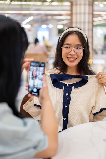 Two happy young women sit in a cafe in a shopping mall, having a fun shopping day together. A woman taking a picture of her friend with a new clothe with a smartphone.