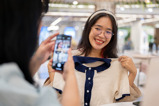 Two happy young women sit in a cafe in a shopping mall, having a fun shopping day together. A woman taking a picture of her friend with a new clothe with a smartphone.