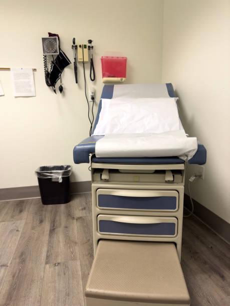 Small medical exam room with inclined table and step. Blue cushion is covered with thin white paper for sanitation. White pillow sits on top. Exam table is beige with step pulled out. Blood pressure cuff and instruments hang on wall