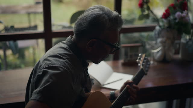 elderly asian man sit by the window and plays an acoustic guitar.