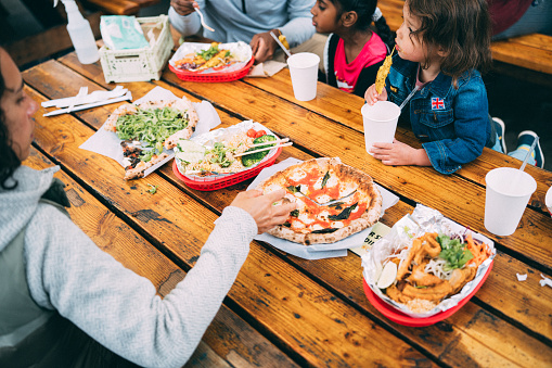 A multiracial group of adults and children gather around a wooden table to enjoy each other's company and eat local cuisine at a food truck pod in Portland, Oregon.