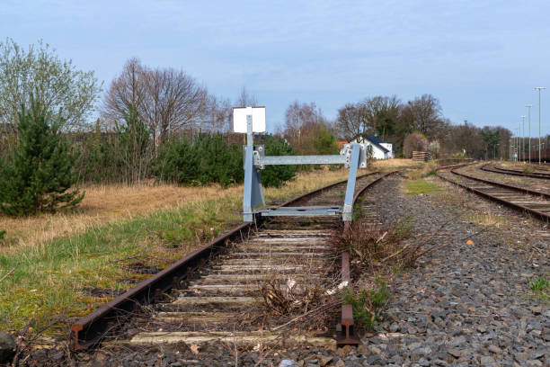 A black metal railway buffer stop located at the end of a set of train tracks. The scene includes surrounding fencing, tracks, and greenery, highlighting an industrial train station setting.