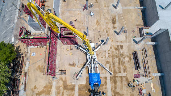 Drone point of view of bright yellow cement pump at construction site, pumping cement into red steel forms for upright walls.