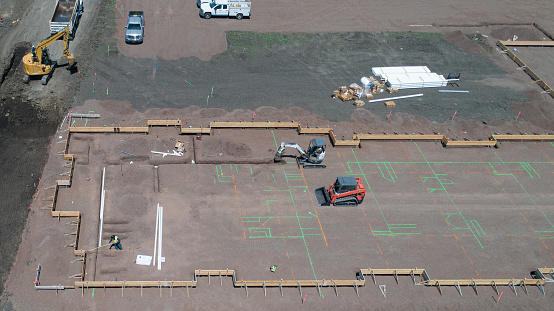 Aerial view of construction site with small digger and scooper following marks on ground to dig trenches for footers and utilities. Beginning phase of construction