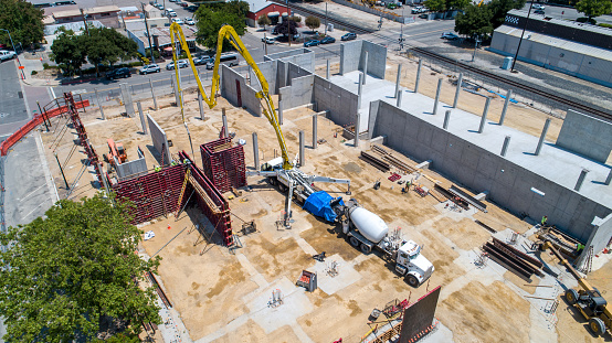 Drone point of view of bright yellow cement pump at construction site, pumping cement into red steel forms for upright walls.