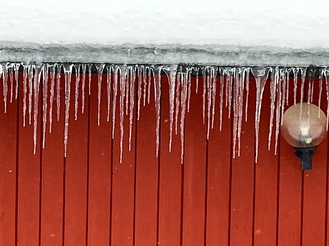 Lots of icicles hanging from snowcovered roof and red wall with a lamp.