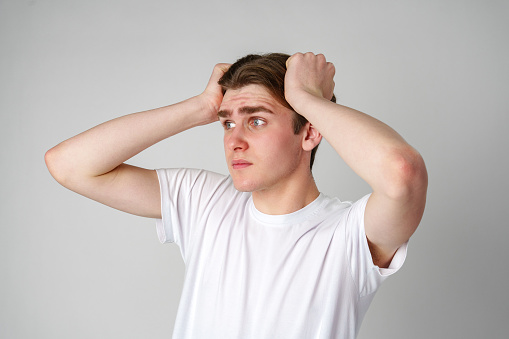 A young man is standing against a plain, light-colored background wearing a simple white t-shirt. He appears worried or stressed, with both hands placed on his head as he looks away from the camera with a furrowed brow.