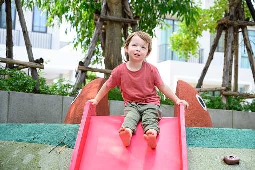 Cute little boy playfull on red plastic slide in playground. Children boy having fun on outdoors playground on sunny summer day. Feeling of fun and enjoyment concept