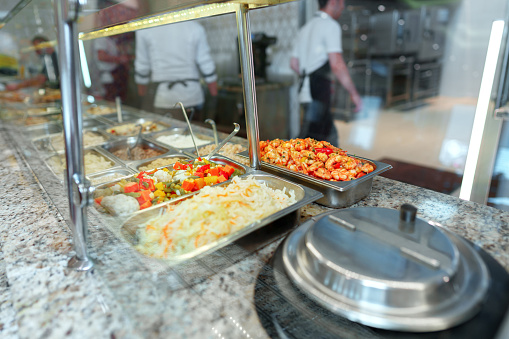 A variety of colorful dishes are neatly presented in a buffet setup with serving utensils. In the background, the blurred figures of attendees and staff suggest an event in progress.