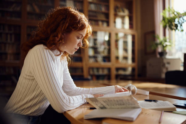 istock Redhead female student reading a book in library. 2147679965