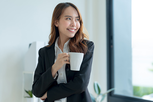 Happy Asian businesswoman in a suit holding a coffee cup Refreshing drink in the office.