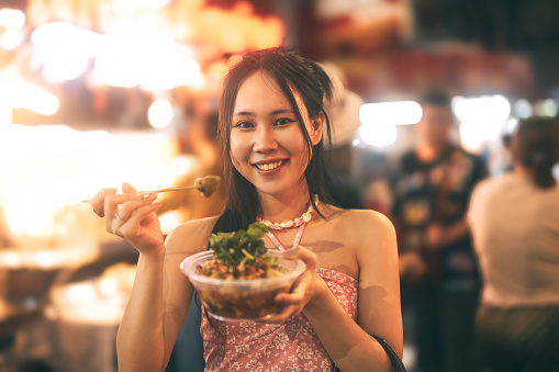 Happy young adult asian tourist foodie woman eating spicy grilled squid. People traveling from tasty guide tour at China town asia street food night market. Bangkok, Thailand