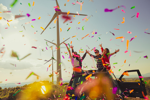 Portrait group of cool teen hipster friends throwing colorful confetti during partying and enjoying the outdoors. They are smiling and looking at the camera posing on a car at a natural wind farm area on a sunny summer day during a carefree road trip. Confetti Joy: Hipster Teens Embrace Carefree Adventure. Capture the vibrant essence of youth as a group of cool and carefree teen hipsters unleashes a burst of colorful confetti during their exhilarating outdoor party. Their infectious smiles and exuberant energy radiate as they pose atop a car at a sun-kissed wind farm area. These friends celebrate friendship, freedom, and the joy of a carefree road trip under the summer sun.