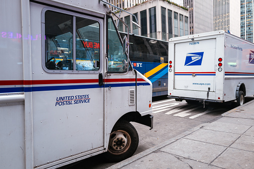 New York, United States of America - February 23, 2024: Wide image of parked USPS vehicles in Manhattan. USPS vehicles, often recognized by their iconic white exteriors and red and blue stripes, are an essential component of the United States Postal Service's fleet. These vehicles, including vans, trucks, and delivery vehicles, crisscross the nation's roads and streets daily, delivering mail and packages to homes and businesses.  The USPS operates a vast fleet of vehicles, including the iconic LLVs, to deliver mail nationwide. In efforts to modernize, newer models like the NGDVs are being introduced for improved efficiency and sustainability. These vehicles symbolize the USPS's commitment to reliable mail service across America.
