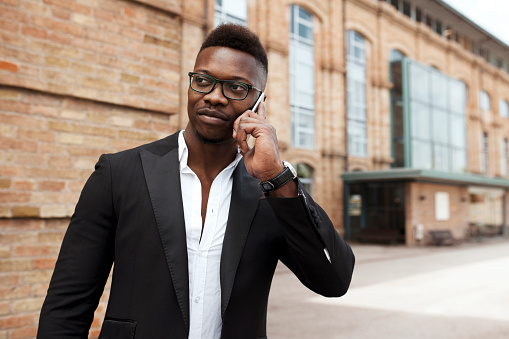 Young stylish african american businessman wears suit and eyeglasses, talking with smartphone standing outdoors at city in front of classic building. Business person, professional, executive, entrepreneur.
