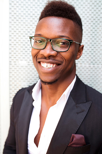 Portrait of young smiling african american businessman wears suit and eyeglasses, standing outdoors at city in front of white wall, looking at camera. Business person, professional, executive, entrepreneur.