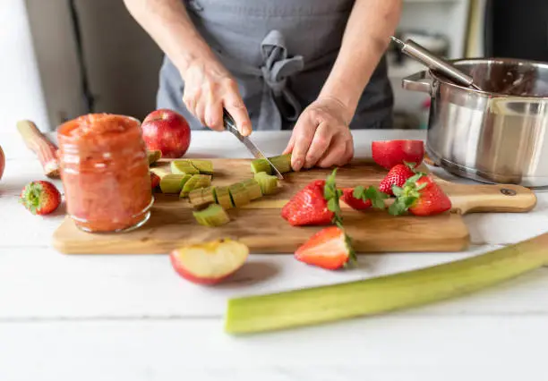 Woman cutting fresh rhubarb on a cutting board for making compote in the kitchen