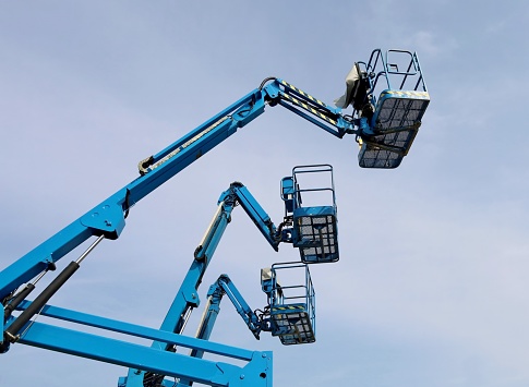 Three blue aerial work platform of cherry pickers, against blue cloudy sky.  Side view.