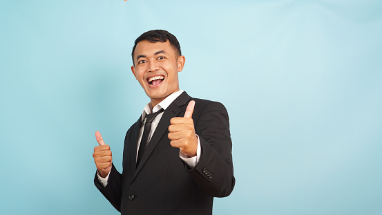 Smiling Asian businessman giving double thumbs up, beaming with success on a blue background