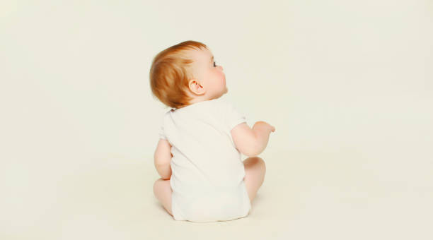 istock Happy cute baby sitting on the floor on white studio background, looking up, back view 2147691122