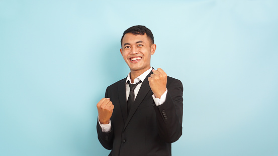 Joyful male executive in a suit with clenched fists, cheerful victory pose, blue backdrop