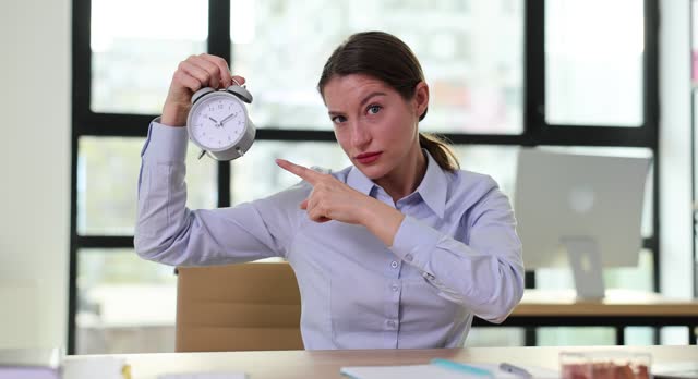 Strict boss businesswoman points to alarm clock at workplace