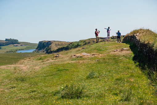 A full shot of a mother and her two adult children hiking a scenic route, crag lough can be seen in the distance. They're all wearing hiking sports attire on a summer's day at Hadrian's Wall, Northumberland. Videos are available similar to this scenario.