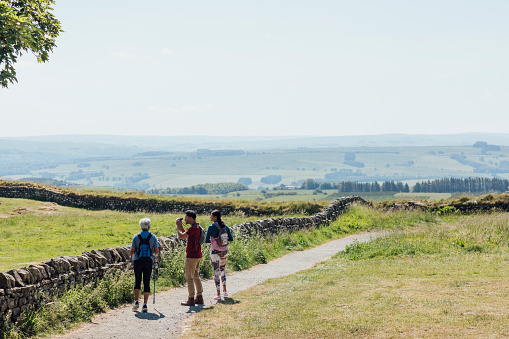 A full shot of a mother and her two adult children hiking a scenic route. They are conversing and the young man is looking through his binoculars. They're all wearing hiking sports attire on a summer's day at Hadrian's Wall, Northumberland. Videos are available similar to this scenario.