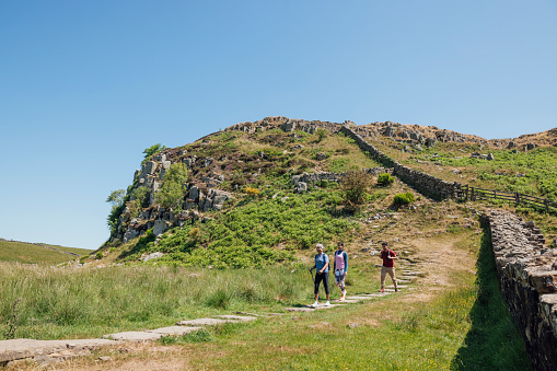 A mother and her two adult children are hiking along a stone pathway engaged in conversation. They're all wearing casual sports attire on a summer's day at Hadrian's Wall, Northumberland. Videos are available similar to this scenario.