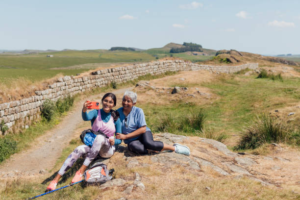 A full shot of Mother and Daughter sitting on a rock in the countryside, they are happily smiling whilst the Daughter holds up her phone to capture a photo. They're both wearing casual sports attire on a summer's day at Hadrian's Wall, Northumberland. Videos are available similar to this scenario.