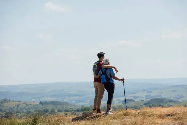 A full shot of a mother with an adult son from behind with a scenic view. The son has his arm wrapped around his mother. They're both wearing casual sports attire on a summer's day at Hadrian's Wall, Northumberland. Videos are available similar to this scenario.