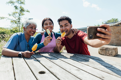 A medium shot of a mother with two adult children sat at a park bench enjoying Ice-creams together and taking selfies. The son is holding the mobile phone. They're all wearing casual sports attire on a summer's day at Hadrian's Wall, Northumberland. Videos are available similar to this scenario.
