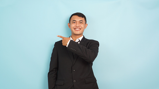 Confident businessman pointing sideways, smiling and presenting, in a sleek black suit, blue background.