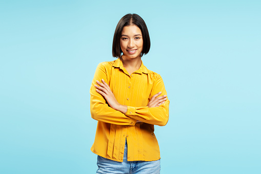 Portrait of smiling beautiful Asian woman, businesswoman wearing casual yellow shirt, arms crossed looking at camera standing isolated on blue background. Business concept, advertising, shopping