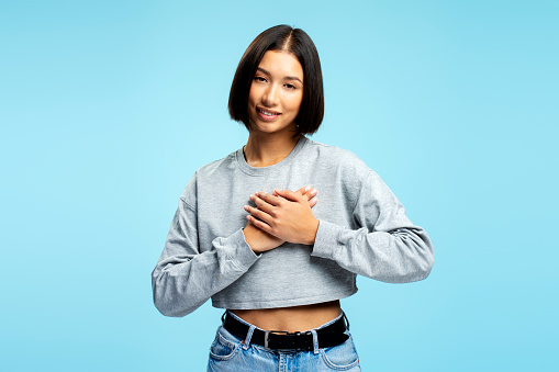 Portrait of young Asian woman with stylish hairstyle holding hands, gesturing, thanking, looking at camera isolated on blue background. Body language concept
