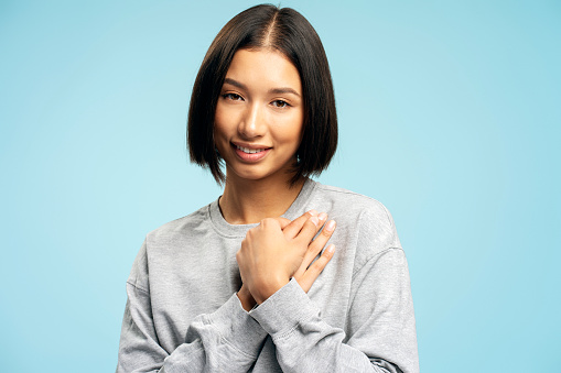 Portrait of smiling Asian woman with stylish bob hairstyle holding hands, gesturing, thanking, looking at camera isolated on blue background. Body language concept
