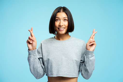 Portrait of smiling young asian woman wearing casual clothes with crossed fingers standing isolated on blue background looking up. Concept of wish something, believing