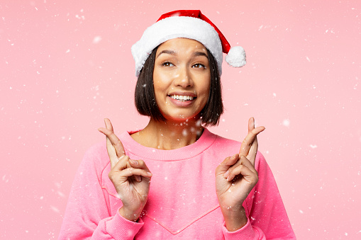 Portrait of smiling beautiful Asian woman wearing red New Year's hat looking away with crossed fingers isolated on pink background, snow falling. Christmas wish concept