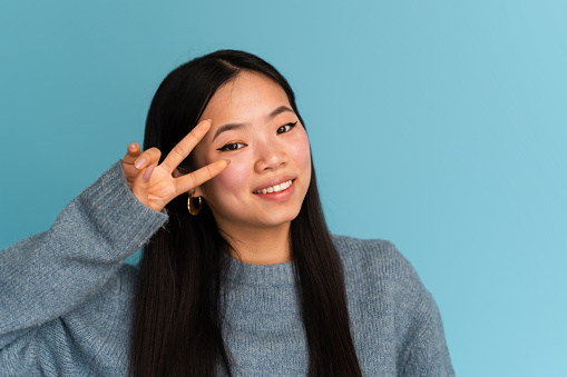 Smiling young woman in a grey sweater gestures peace against a pastel blue backdrop