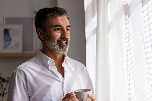 Smiling bearded man in a white shirt holding a coffee cup by the window with soft light
