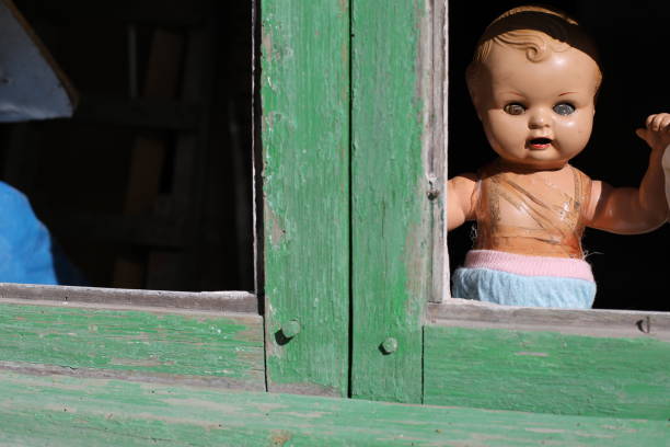 A spooky porcelain baby doll is looking through a window that seems to belong to a secret and abandoned place in the middle of nowhere. The setting is perfect for a Halloween decoration