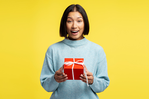 Portrait of smiling beautiful Asian woman wearing casual sweater, holding red gift box and looking at camera. Conception Celebrations, Birthday