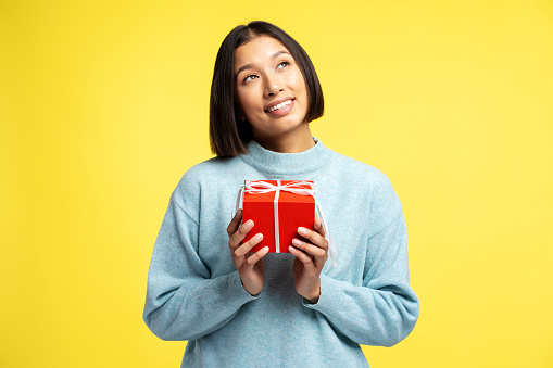 Portrait of smiling Asian woman dreaming holding red gift box looking up standing isolated on yellow background. Concept of celebration, holidays, birthday