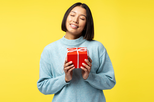Positive cheerful Asian woman wearing casual sweater holding red gift box with closed eyes standing isolated on yellow background. Concept of celebration, holidays, birthday